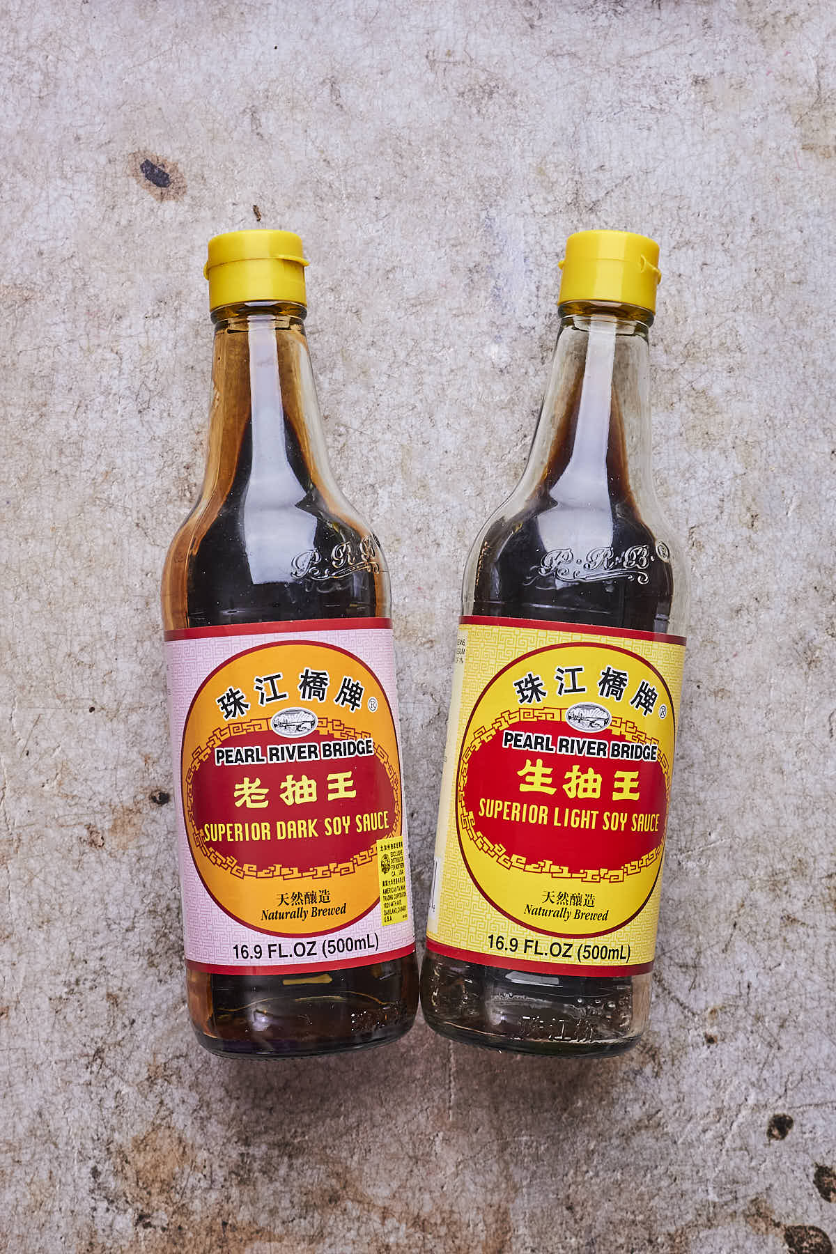 Two bottles of Pearl River Bridge soy sauce, one dark and one light, on a textured gray surface.