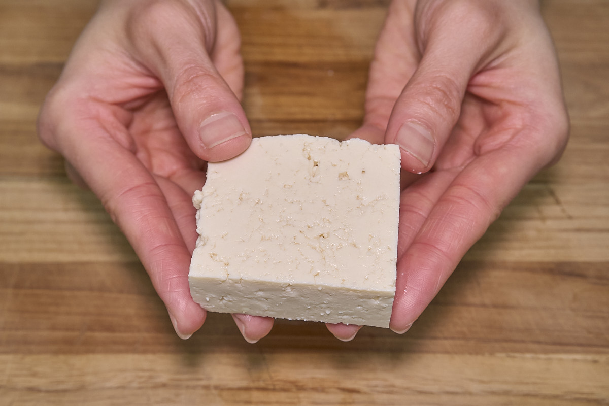 Two hands holding a block of tofu over a wooden surface.